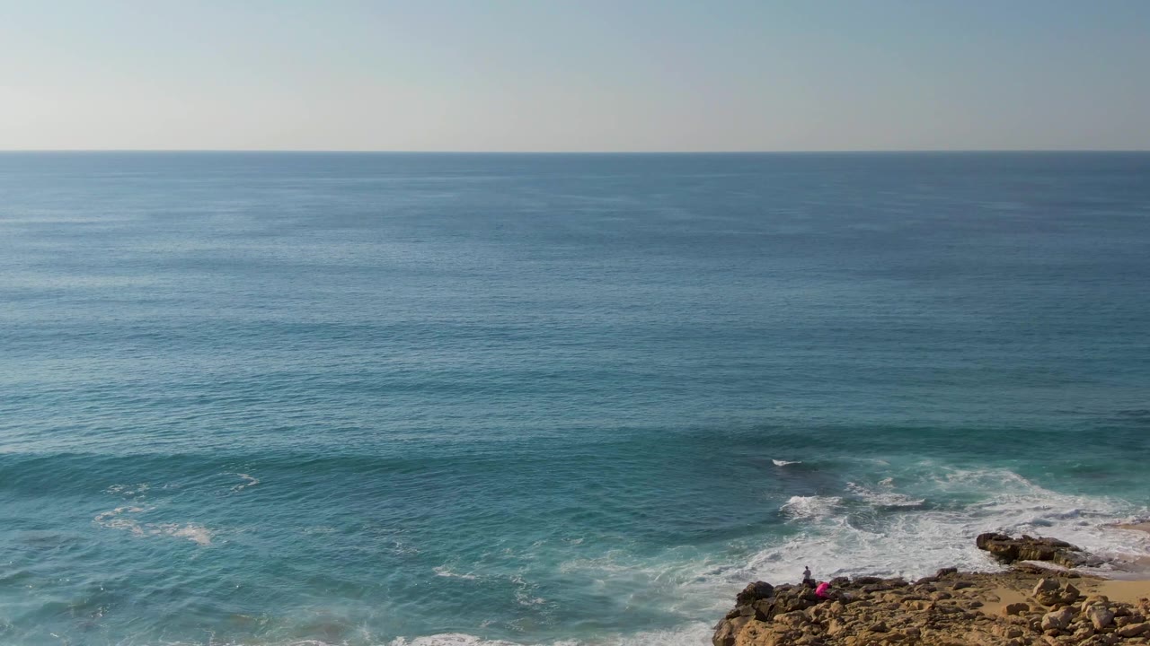 Turquoise water and white surf break against rocky shore at Buzzards Beach East Cape BCS Mexico, with a small person in pink visible near the rocks at the lower right.