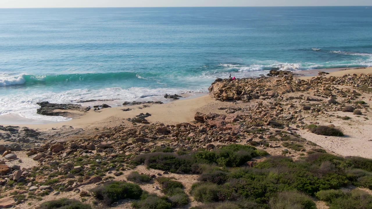 Wide aerial coastline with waves, rocks, sand, and green scrub