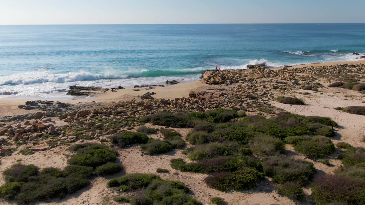 Aerial frame at Buzzards Beach East Cape BCS Mexico showing blue ocean, white surf, rocky sand, low shrubs in the foreground, and a tiny distant person on the rocks.