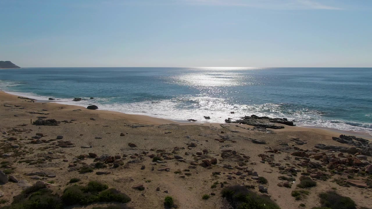 A second aerial moment over Buzzards Beach East Cape BCS Mexico shows the shoreline angled across frame, with beach rocks below, foamy waves midframe, sun reflection on the sea, and no visible people.