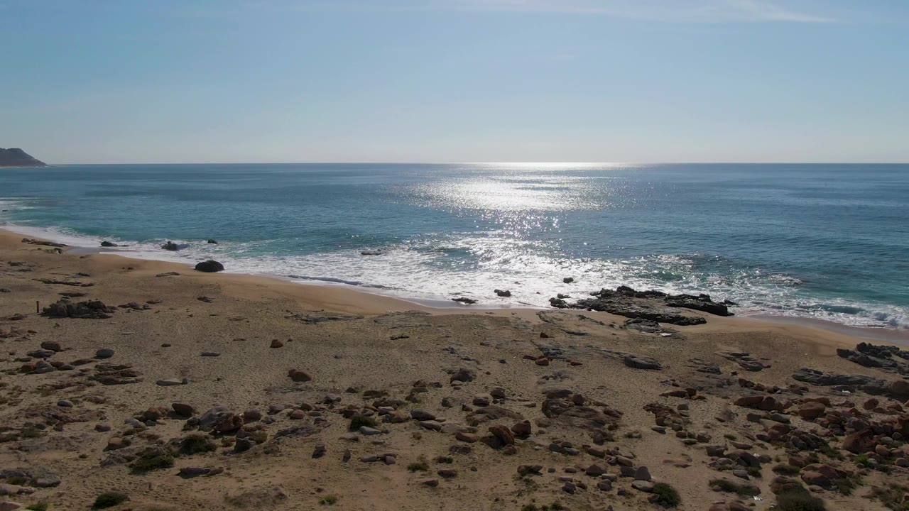 Buzzards Beach East Cape BCS Mexico appears from a high angle with fewer shrubs in view, more open sand, a rocky shoreline, white wave foam, strong sun glint, and no visible people.