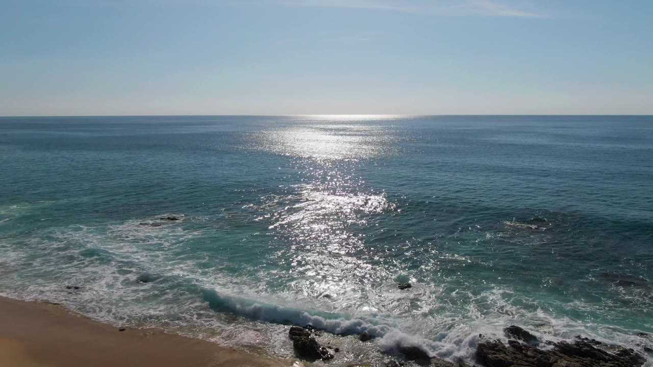 Buzzards Beach East Cape BCS Mexico frame with a narrow strip of sand, dark rock formations, active white surf, broad blue water, sun reflection, and no visible people.