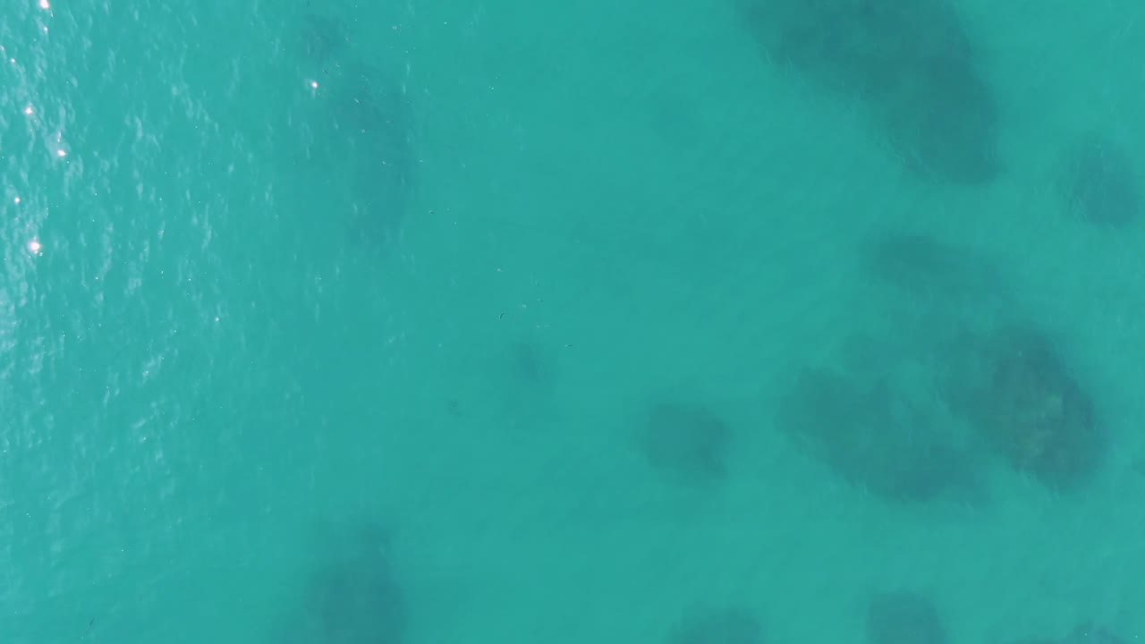 Aerial frame of blue-green shallow ocean at Buzzards Beach, East Cape, BCS, Mexico, with several dark underwater patches softened by rippled water and no visible people.