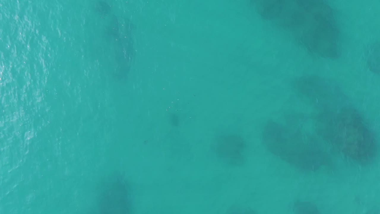 Top-down turquoise ocean frame from Buzzards Beach, East Cape, BCS, Mexico, showing broader dark reef or rock shapes beneath a lightly rippled surface and no visible people.