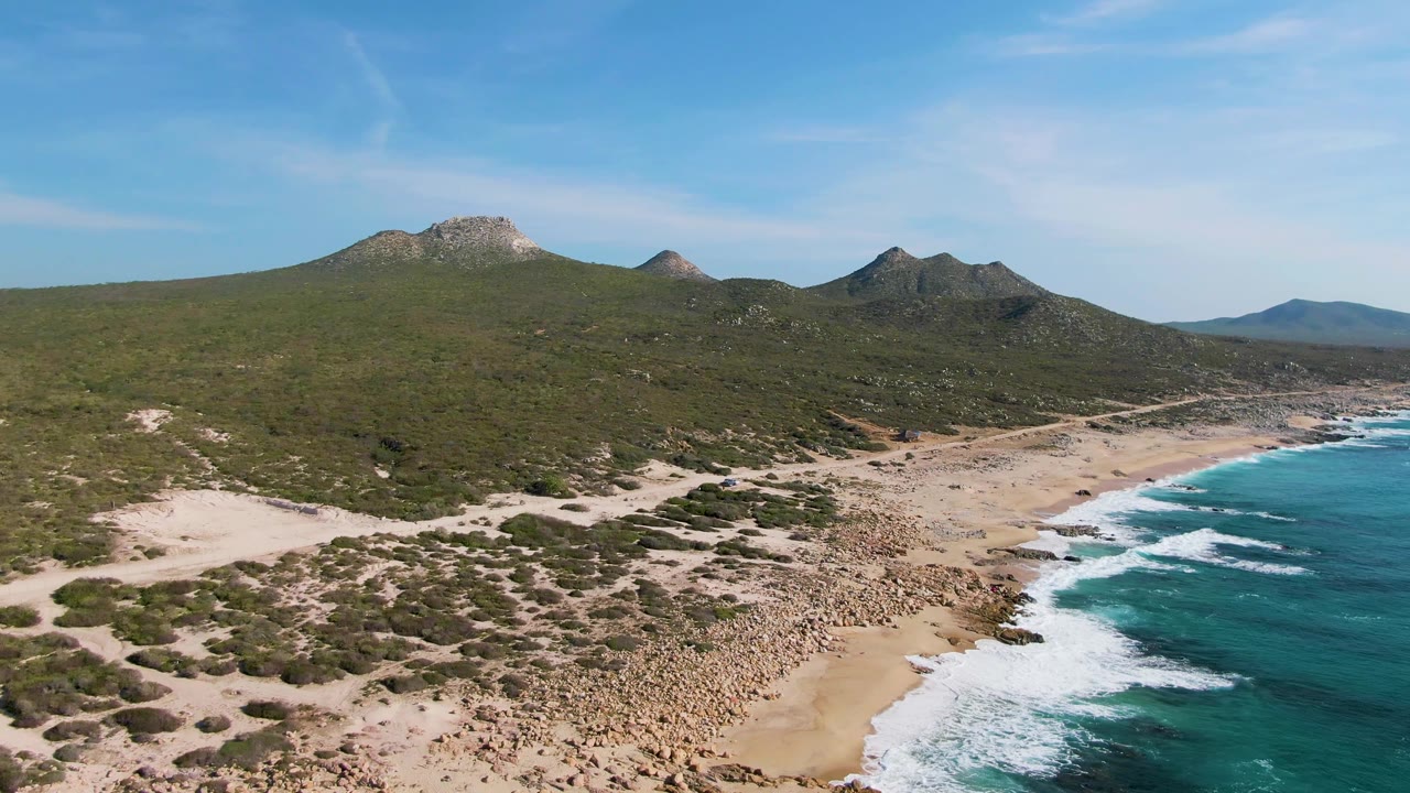 Opening aerial frame over Buzzards Beach in East Cape BCS Mexico, showing pale sand, rocky shore, turquoise waves, scrub-covered hills, and no visible people.