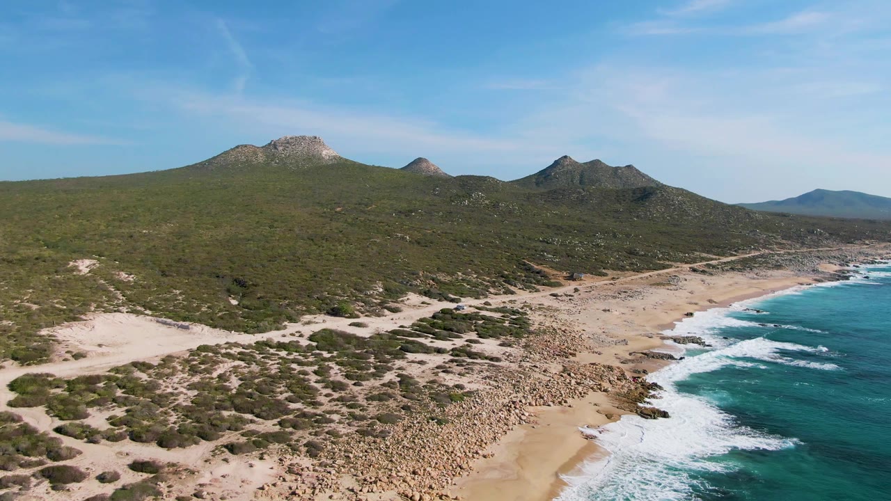 Drone view along a sandy shoreline with waves and shrub-covered hills.