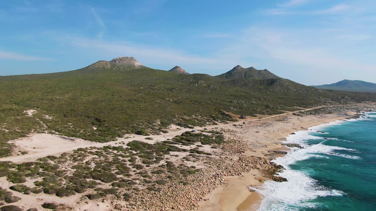 Two seconds in, the Buzzards Beach aerial shows a broader rocky section of sand, bright surf at frame right, low green vegetation, distant hills, and no visible people.