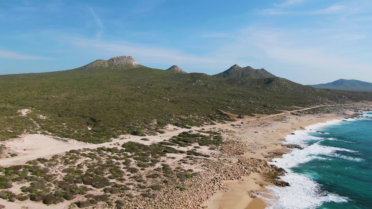 Coastal aerial with sandy beach, waves, scrubland, and rounded hills.
