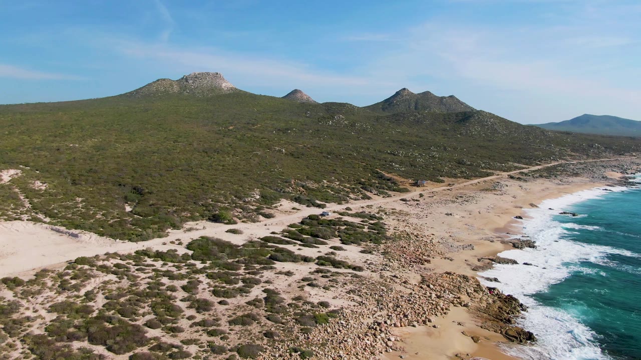 Aerial view at Buzzards Beach in Mexico showing scrubby dunes, a rocky sandy shoreline, green mountain slopes, blue sky, and white surf at the right edge, with no visible people.