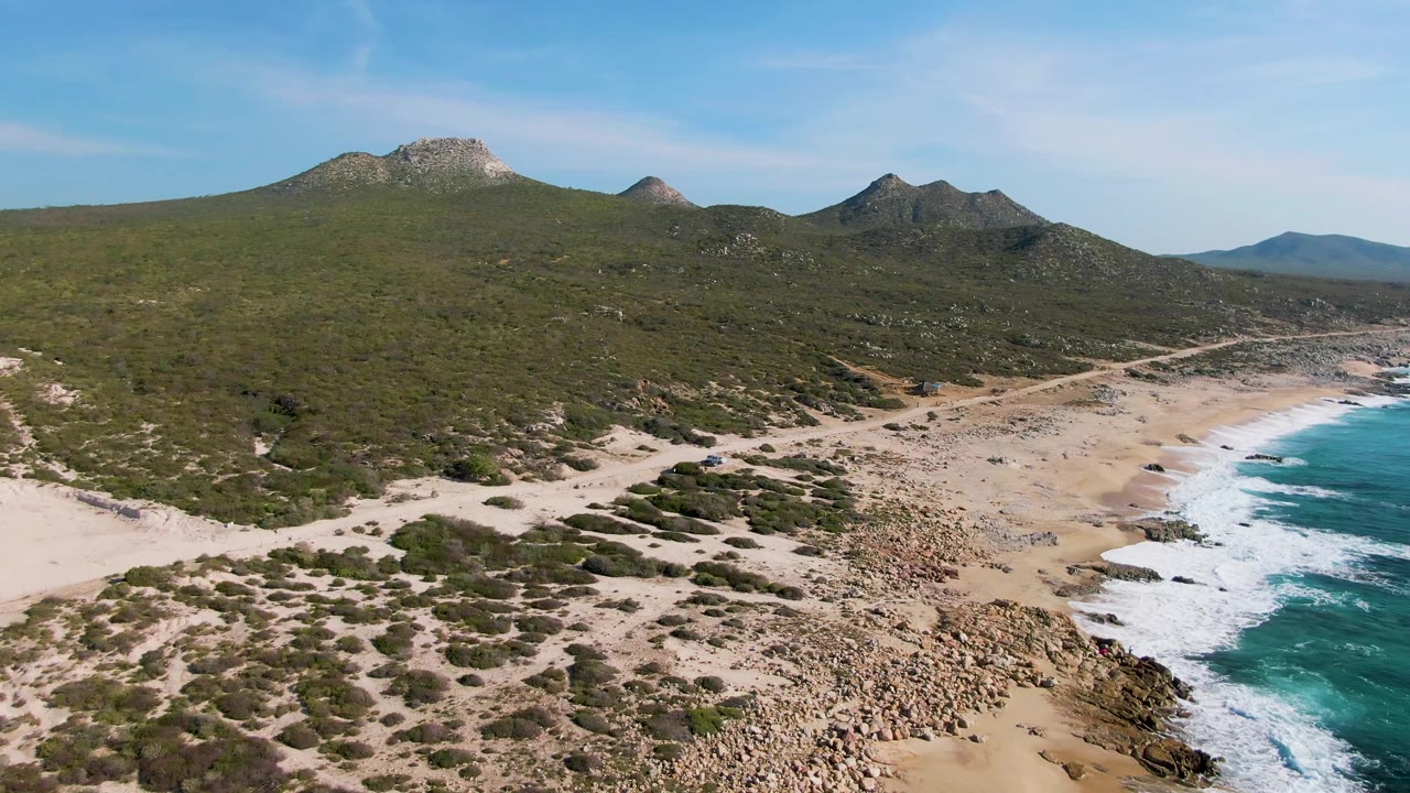 Wide aerial frame at Buzzards Beach in Mexico with green hills filling the upper half, a pale sandy beach and rock field below, and turquoise water breaking into white foam on the right, with no visible people.