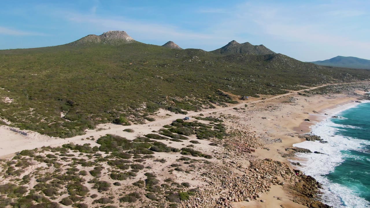 Aerial view over Buzzards Beach in Mexico showing a broad green hillside, multiple rocky peaks, pale sand, scattered shoreline rocks, and a band of ocean surf at frame right, with no visible people.