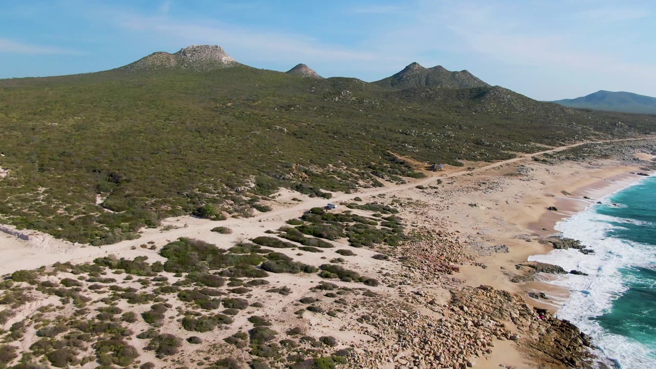 Elevated beach view with scrubby foreground, rocky shore, green hills, and waves.