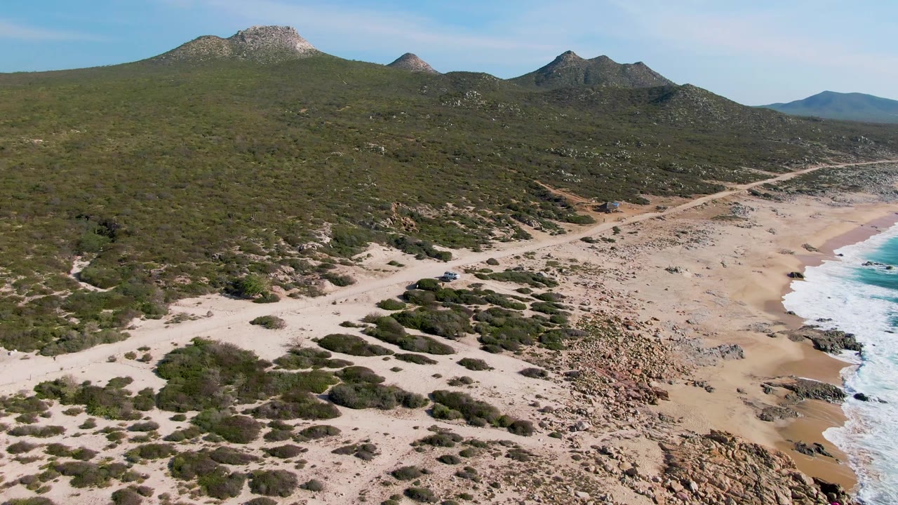 Aerial view of a dirt road beside a sandy beach and rocky surf line.