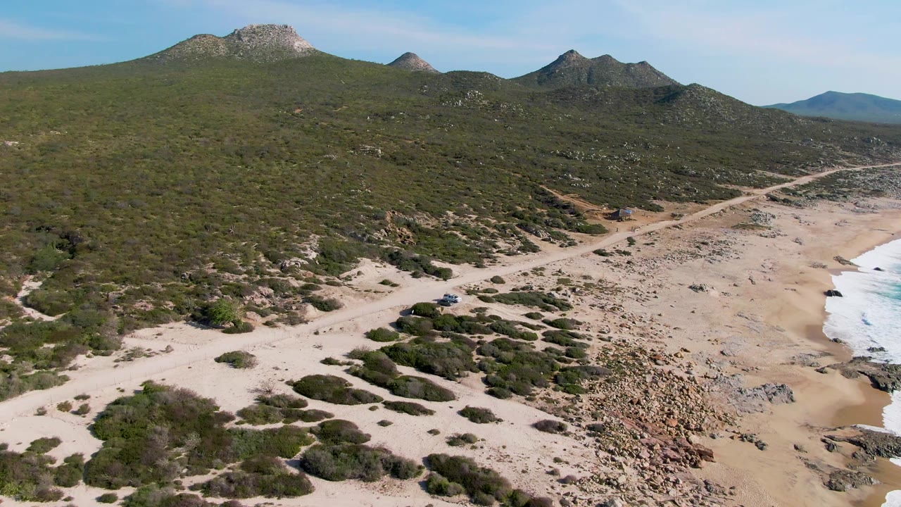 Elevated frame over Buzzards Beach East Cape BCS Mexico showing the dirt road tracking along the beach, low vegetation spreading inland, hills on the horizon, and no visible people.