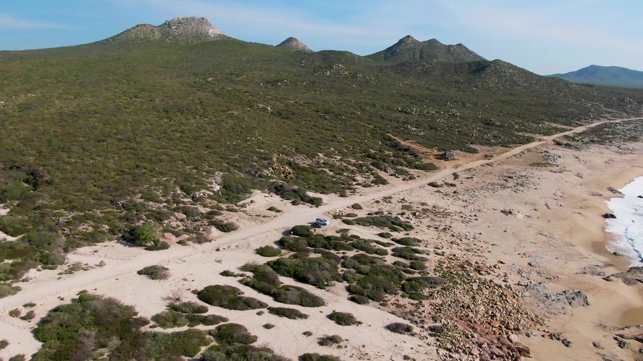 Aerial view at Buzzards Beach East Cape BCS Mexico with scrub-covered hills filling more of the frame, the dirt road crossing the lower beach area, surf at right, and no visible people.