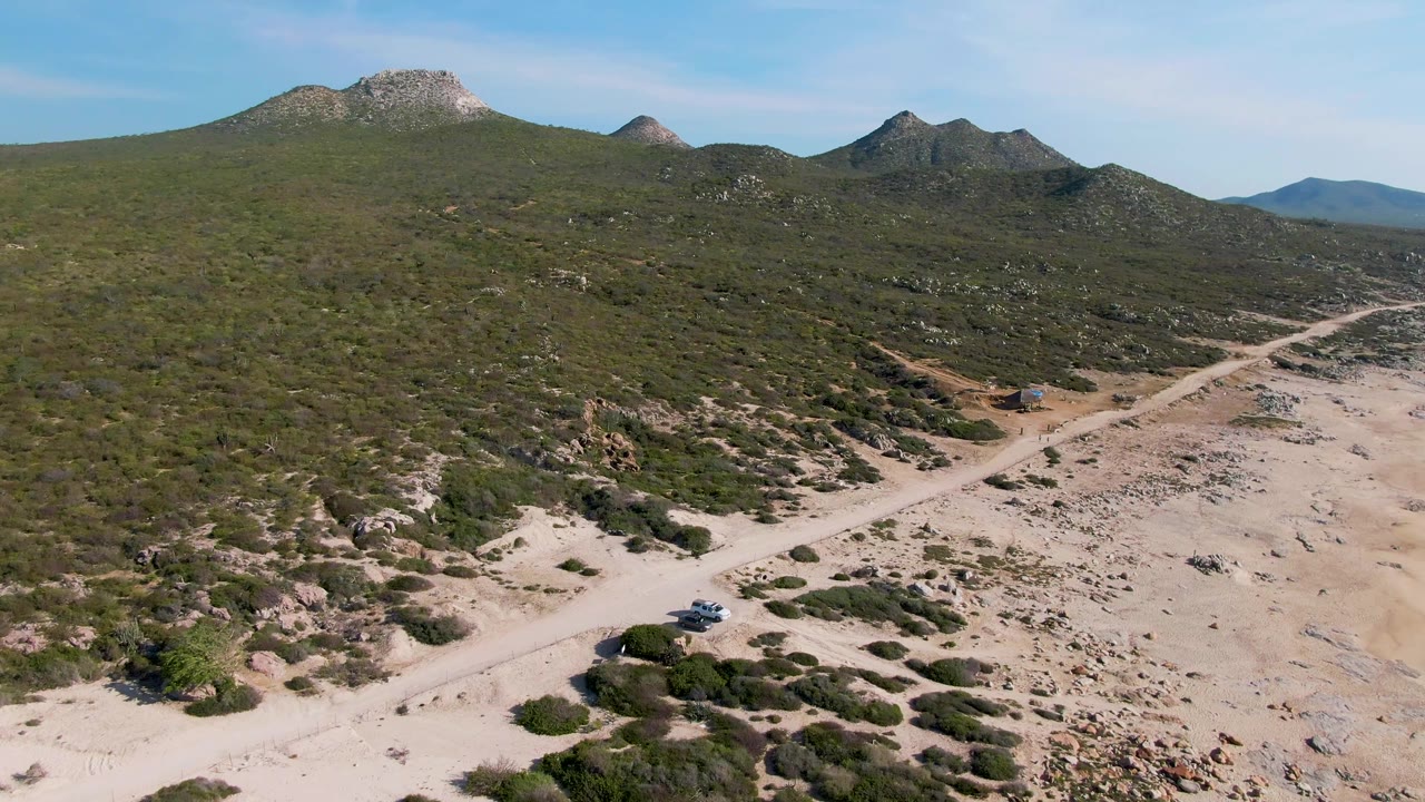 Car on a dirt road below scrub-covered hills and rocky mountains.
