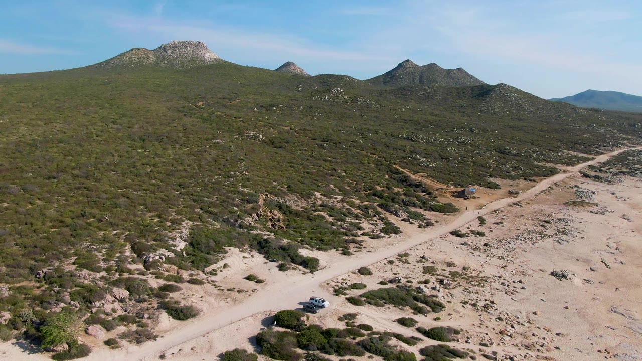 Aerial frame from Buzzards Beach East Cape BCS Mexico showing the car near a pale forked dirt road, scattered desert vegetation, rocky hillsides, mountain peaks, and no visible people.