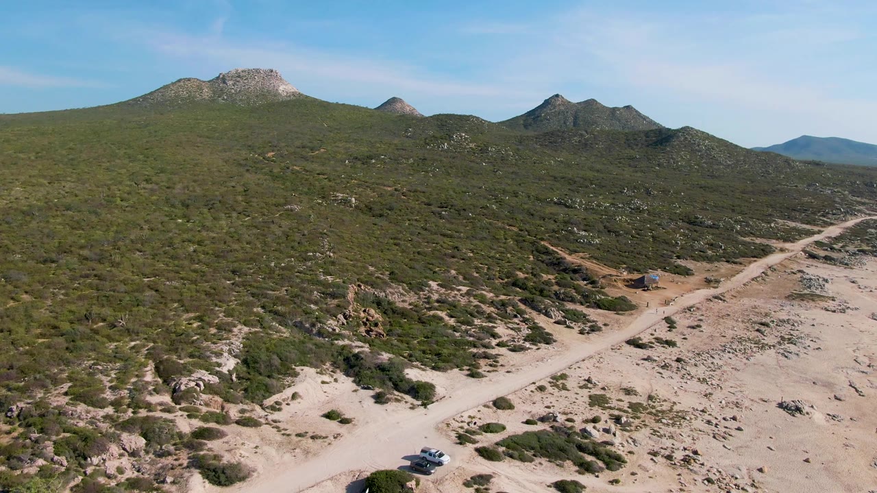 Aerial view in Buzzards Beach East Cape BCS Mexico with the car lower in frame on a dusty road, rocky scrubland rising behind it, low mountain peaks, and no visible people.