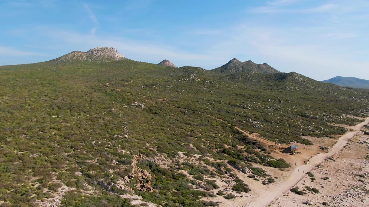 Aerial frame from Buzzards Beach East Cape BCS Mexico showing a pale dirt road, low green shrubs, rocky hills on the horizon, and no visible people.