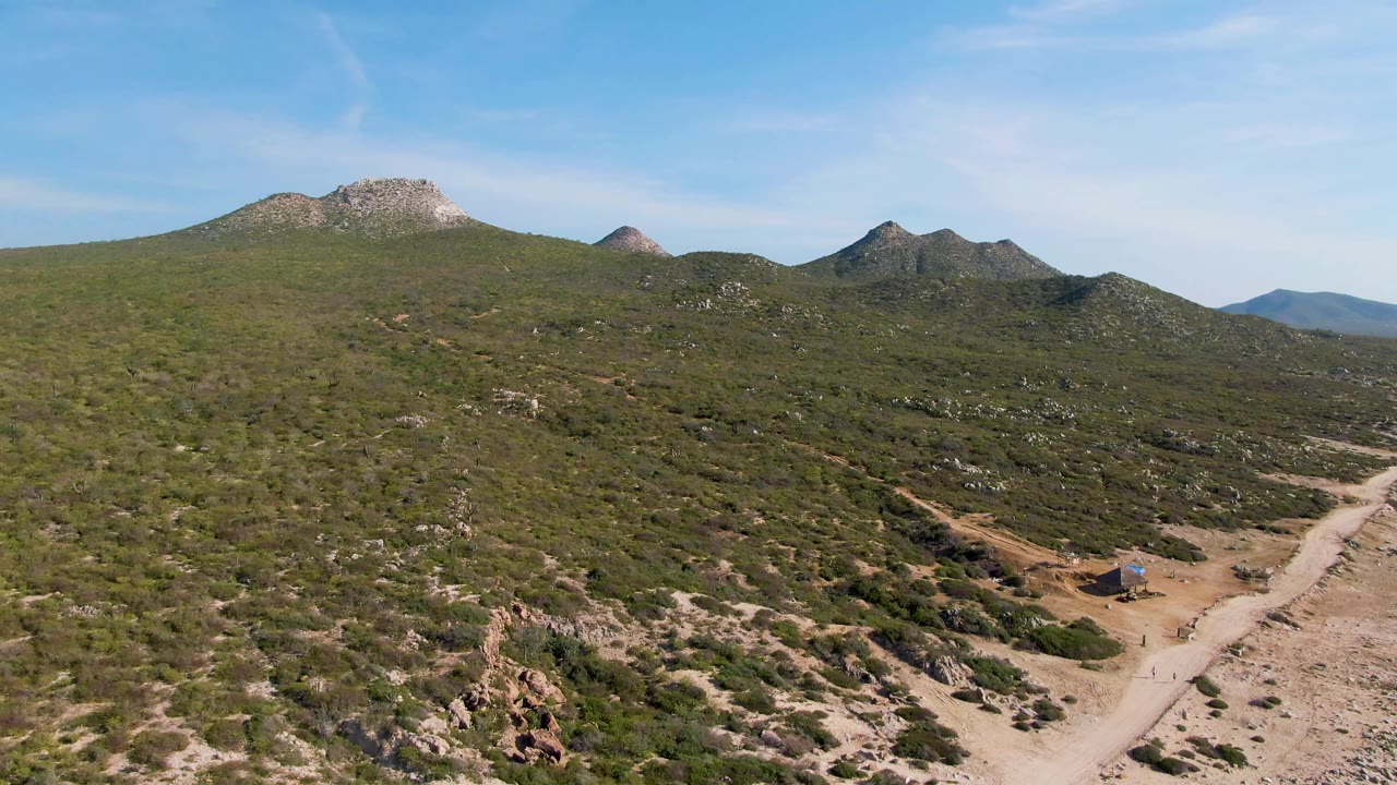 Aerial view in Mexico with the dirt road more centered through scrubby terrain, a small vehicle or structure near the road, rocky ridges beyond, and no visible people.