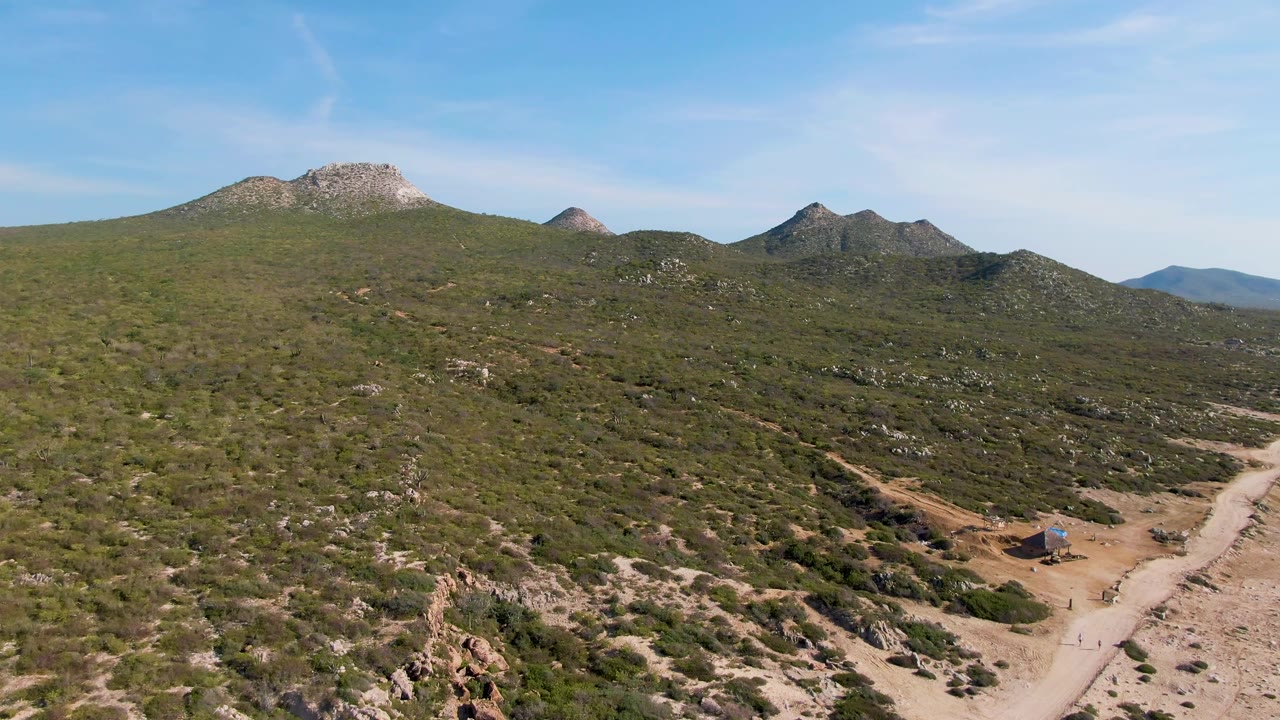 Wide aerial frame from the East Cape clip showing shrubland, a diagonal dirt road, rocky hilltops across the background, blue sky, and no visible people.