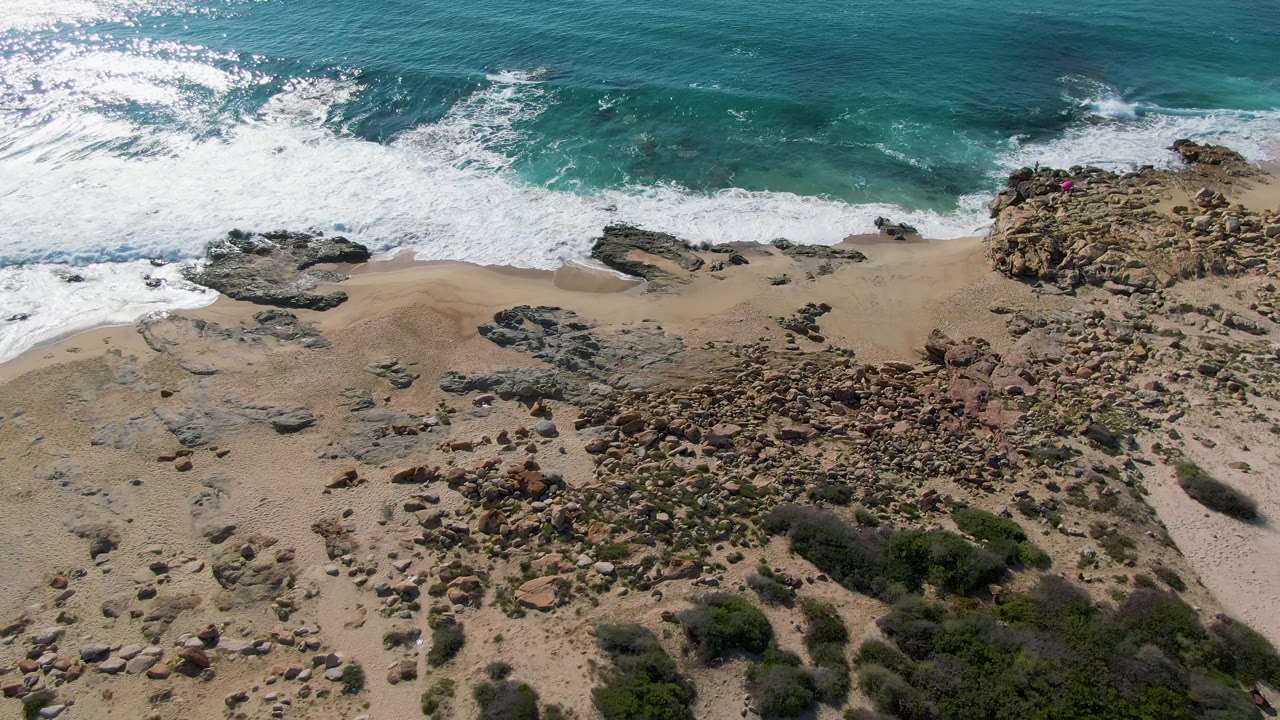 Aerial frame over Buzzards Beach in Mexico showing white surf breaking along turquoise water, rocky sand, scrub vegetation, and no visible people.