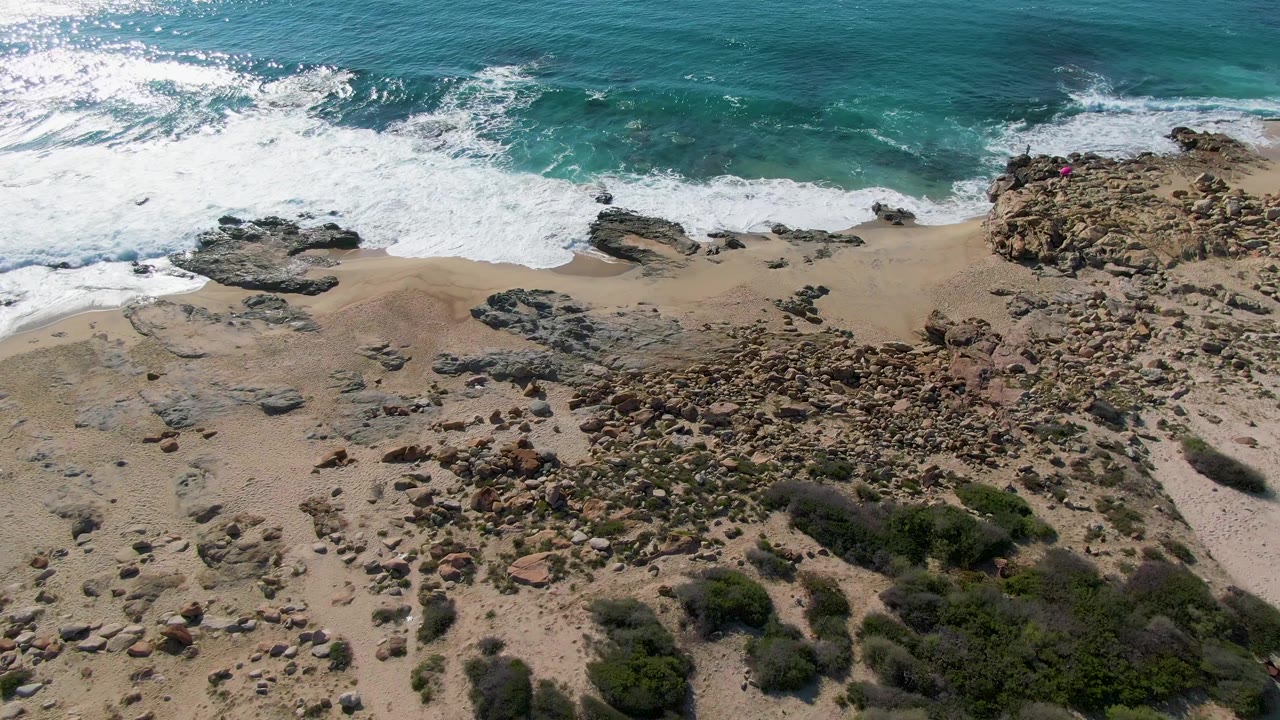 Aerial frame over Buzzards Beach in Mexico with white water spreading across shore rocks, pale sand below, green scrub near the bottom edge, and no visible people.