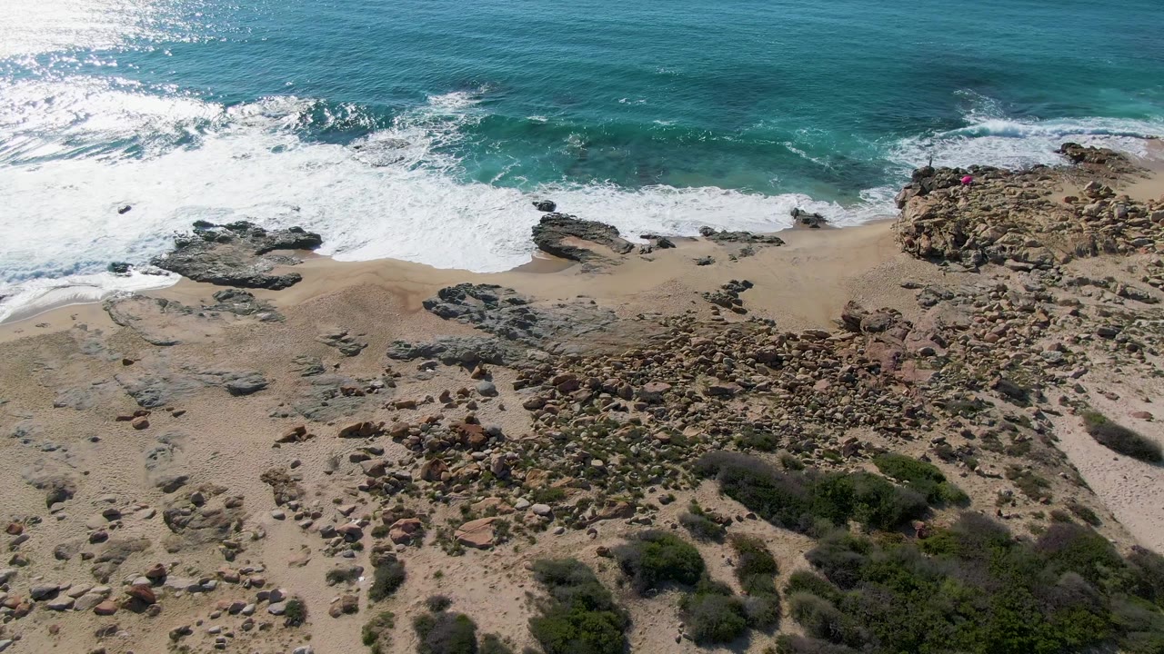 Aerial frame over Buzzards Beach in Mexico showing a broad rocky section of beach beneath breaking turquoise surf, with dry vegetation and no visible people.