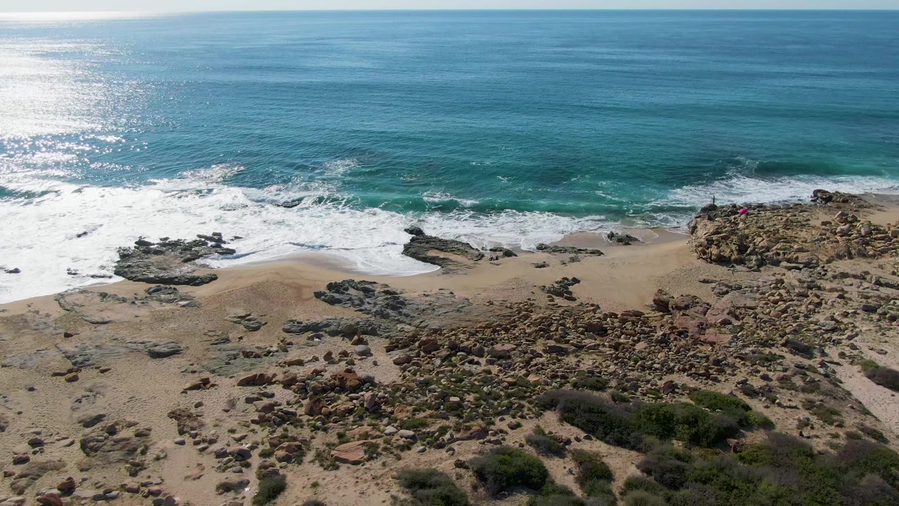 Aerial frame over Buzzards Beach East Cape BCS Mexico with sunlit ocean, white surf, sandy shoreline, rocky outcrops, scrub vegetation, and a tiny possible person on the right-side rocks.