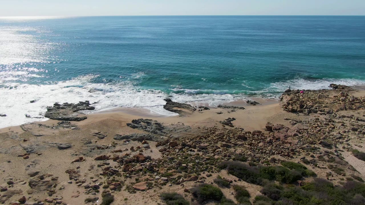 Drone view at Buzzards Beach East Cape BCS Mexico with white waves spreading across sand, dark flat rocks in the surf zone, blue water beyond, scrub foreground, and no visible people.