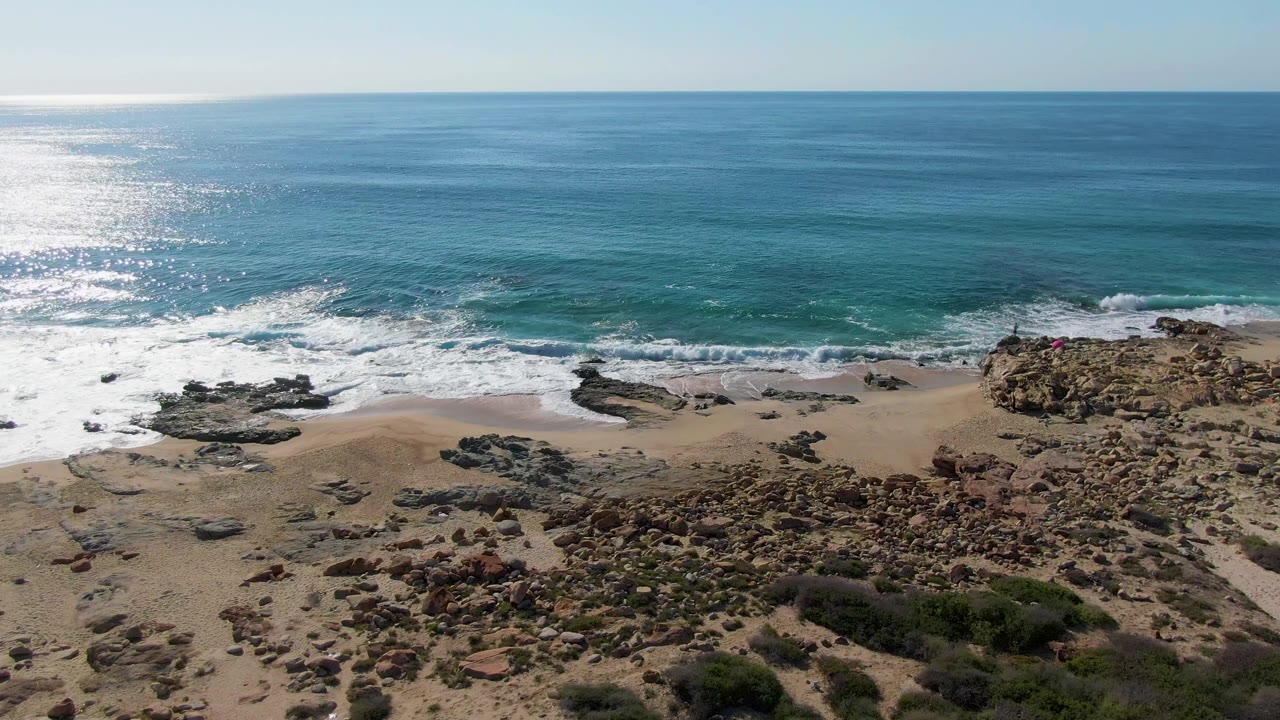 Aerial view of Buzzards Beach East Cape BCS Mexico with turquoise water near the beach, white foam curling along the sand, rocky ground at right, sparse vegetation below, and no visible people.