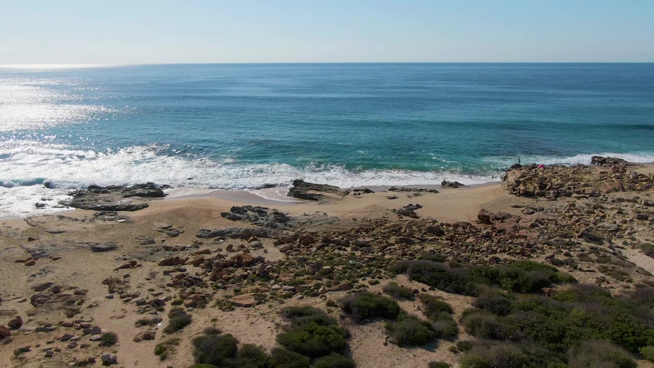 Aerial frame from Buzzards Beach East Cape BCS Mexico showing blue ocean, white surf, dark rock shelves, sandy shore, foreground scrub vegetation, and no visible people.