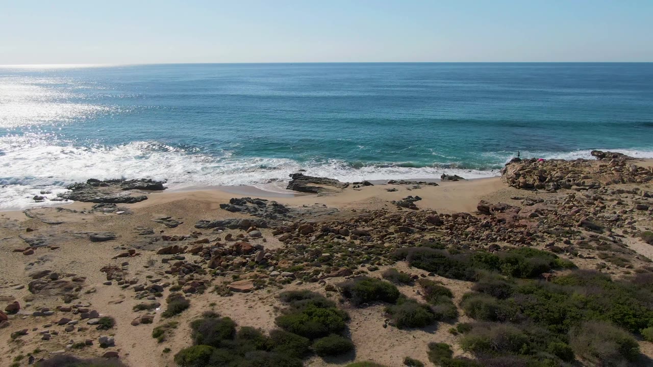 Aerial frame from Buzzards Beach East Cape BCS Mexico with bright sun glare on the left water, a fresh line of foam at the shore, scattered rocks, low green shrubs, and no visible people.