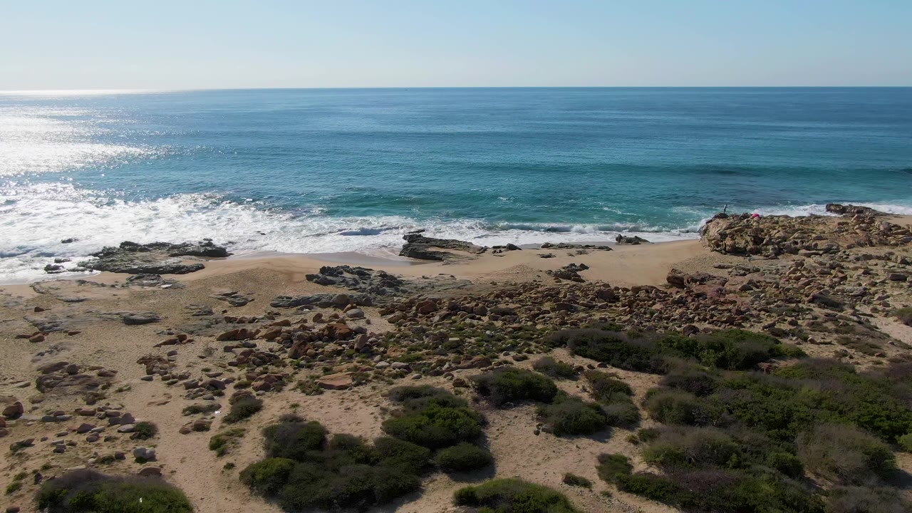 Aerial frame from Buzzards Beach East Cape BCS Mexico showing a wider spread of scrub in the foreground, white water along the beach, turquoise shallows, darker offshore water, and no visible people.