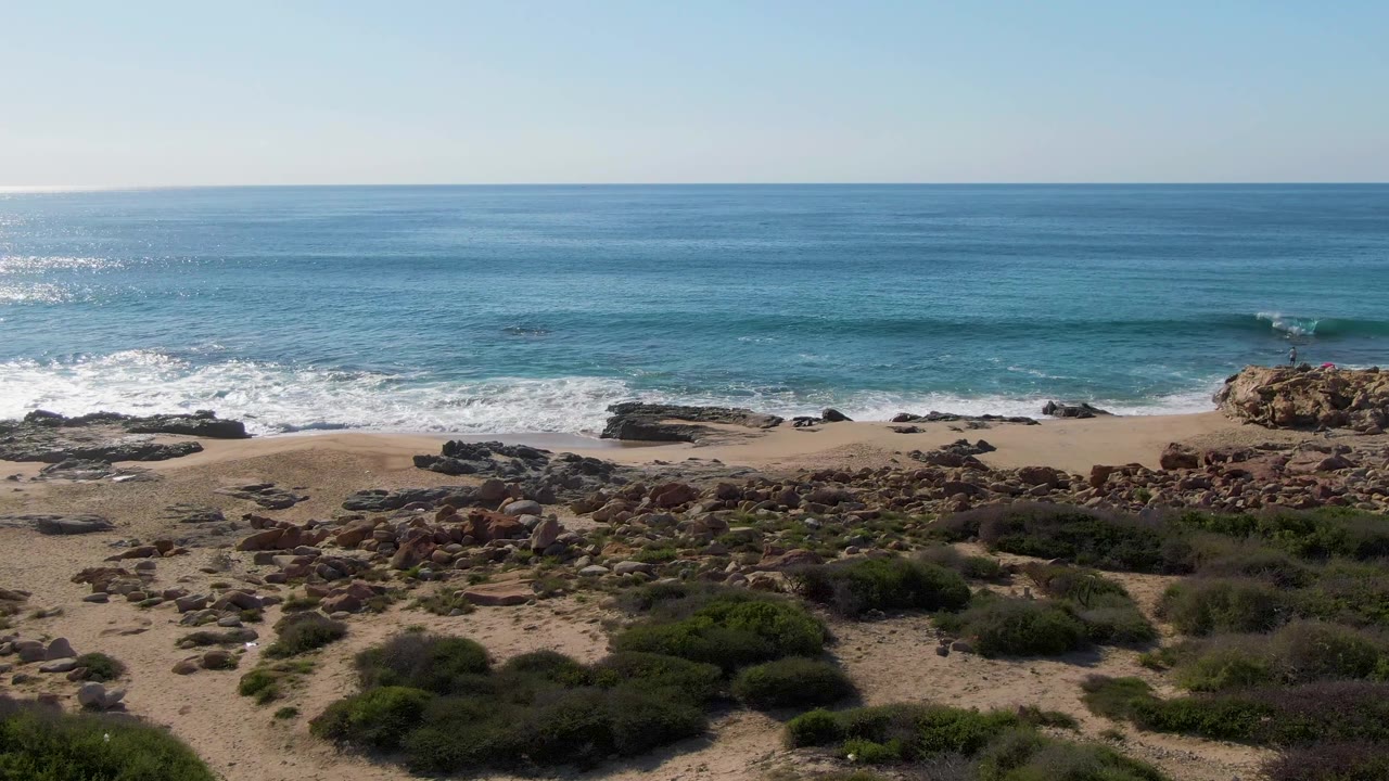 At Buzzards Beach in Mexico, a line of white foam spreads along the sand while dark rocks and green shrubs fill the lower shore, with no visible people.