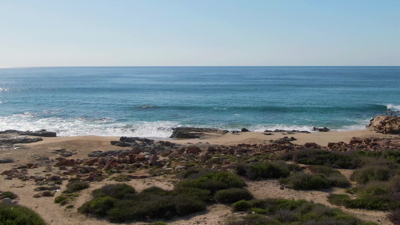 At Buzzards Beach in Mexico, foreground shrubs and scattered rocks lead toward a sandy strip where white waves meet the shore, with no visible people.