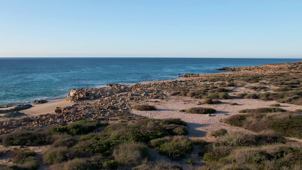 Opening frame of Buzzards Beach East Cape BCS Mexico with rocky shoreline, pale sand, low dune vegetation, blue ocean, clear horizon, and no visible people.