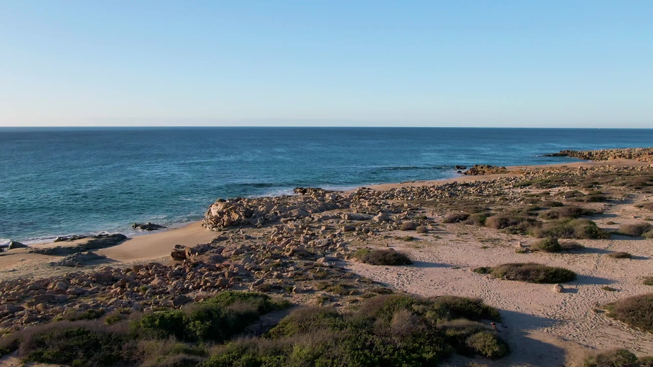 Opening frame from Buzzards Beach East Cape in Mexico showing blue ocean beyond a rocky shoreline, pale sand, low scrub vegetation, clear sky, and no visible people.