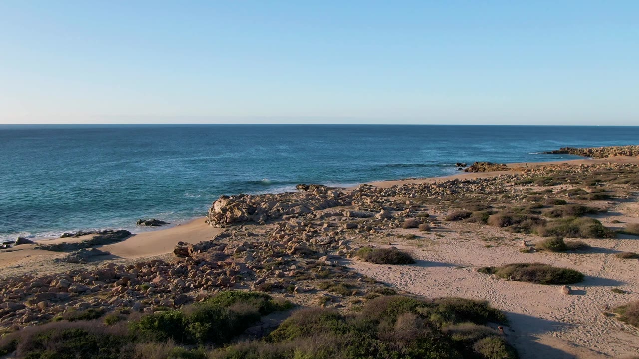 At one second, Buzzards Beach East Cape appears from a slightly advanced aerial angle, with small waves nearing dark rocks, a sandy pocket beach, scrub-covered ground, and no visible people.
