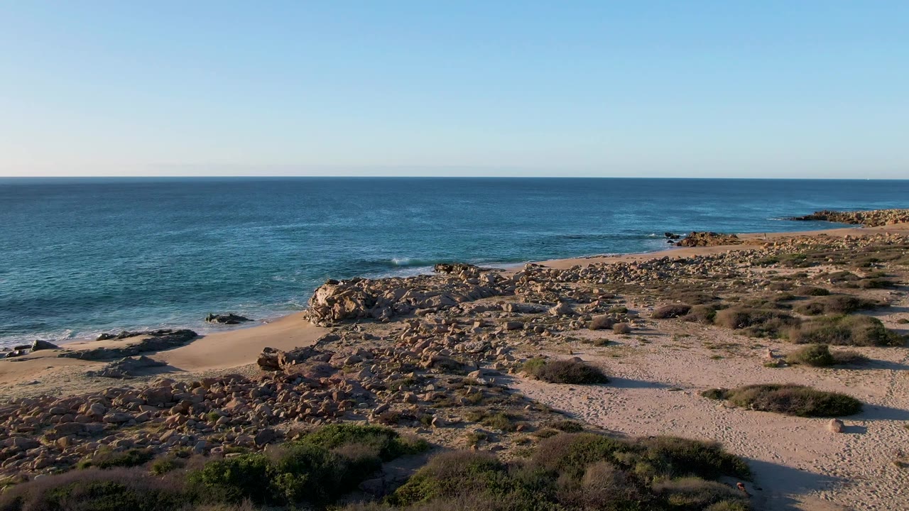 At two seconds, the aerial view holds on Buzzards Beach East Cape with a rocky point across the midground, blue sea behind it, sandy paths or open sand below, and no visible people.