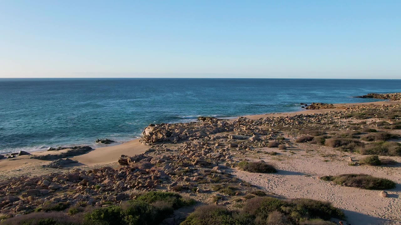 Wide beach and rocky shoreline with gentle surf under a clear sky.