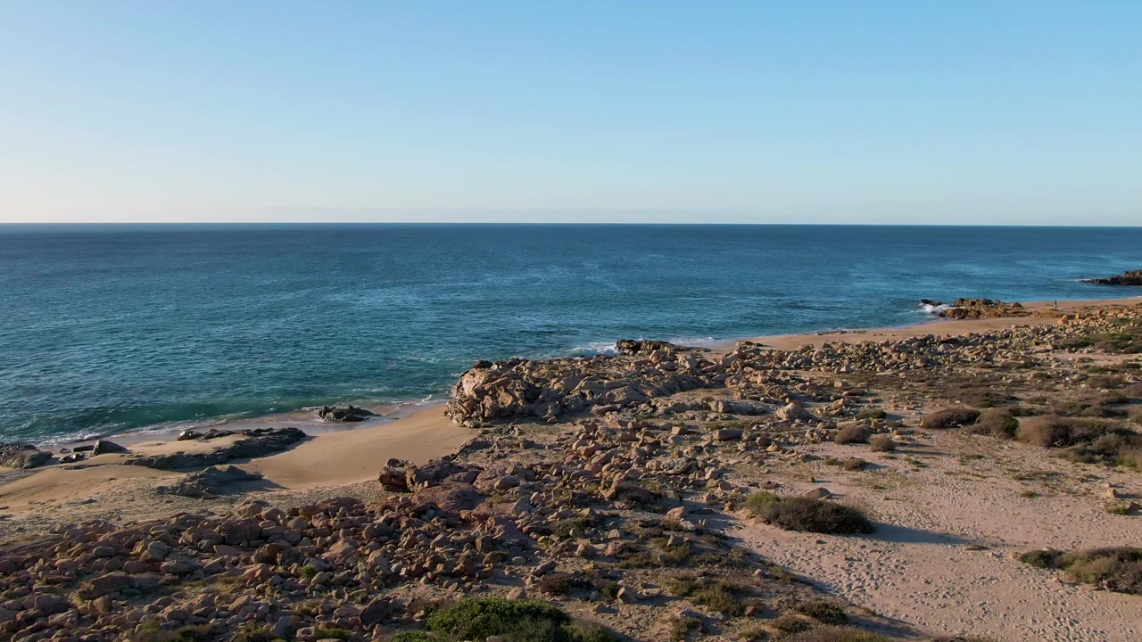 Opening frame of Buzzards Beach East Cape BCS Mexico with tan rocks in the foreground, a sandy cove at left, blue ocean beyond, and no visible people.