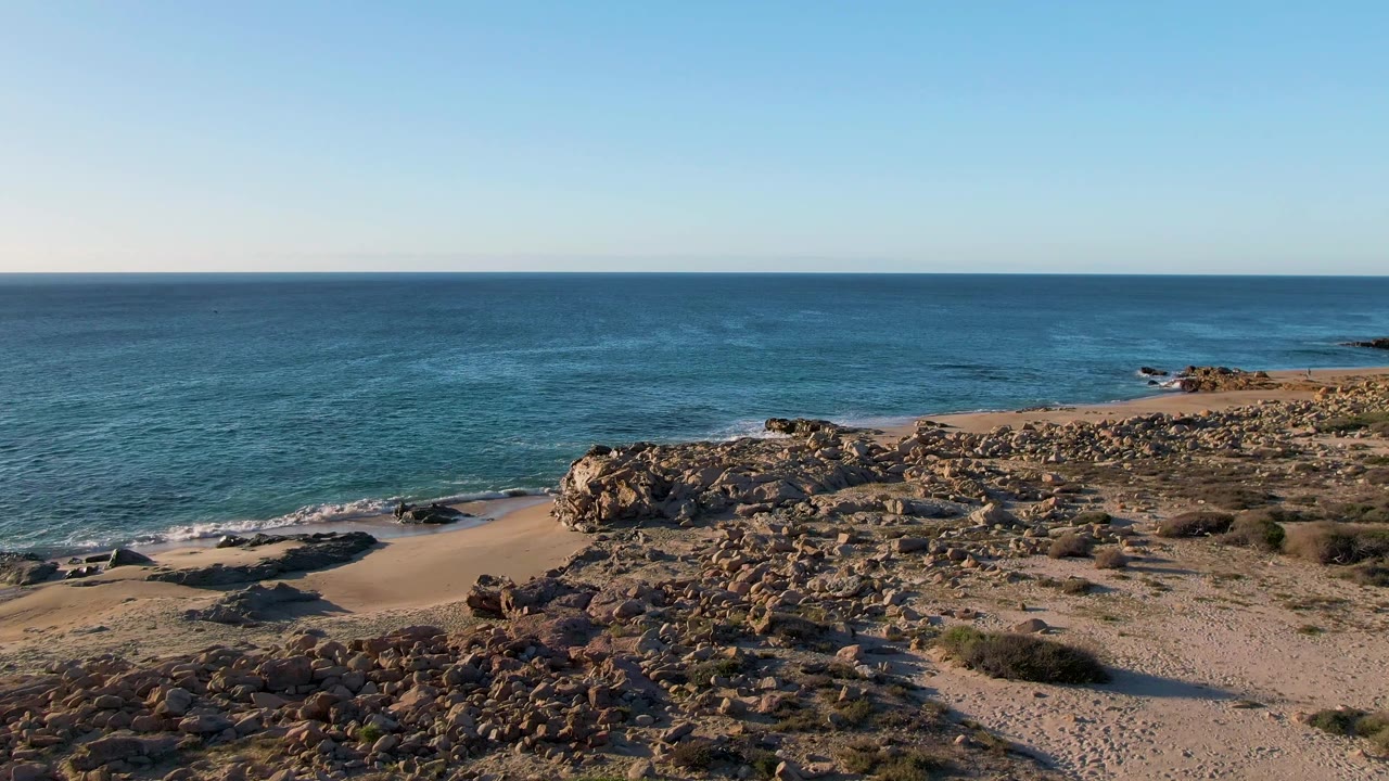 A later frame shows Buzzards Beach East Cape BCS Mexico with small white wave edges at the left beach, rocky outcrops across the center, and no visible people.