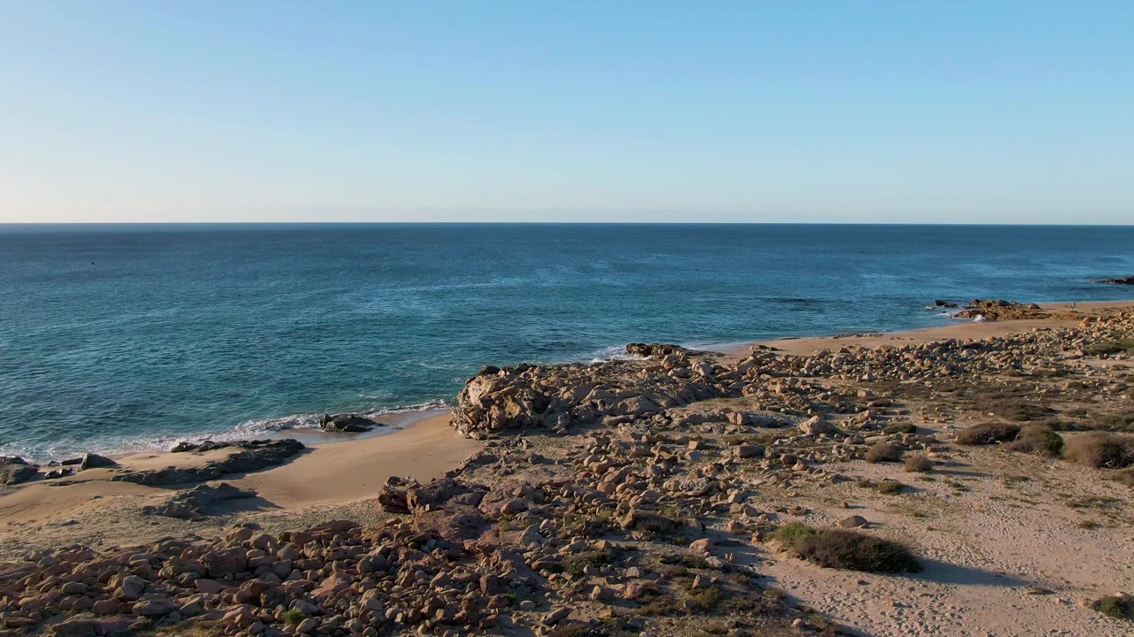 Open ocean beyond a rocky, sandy shoreline under clear daytime sky.