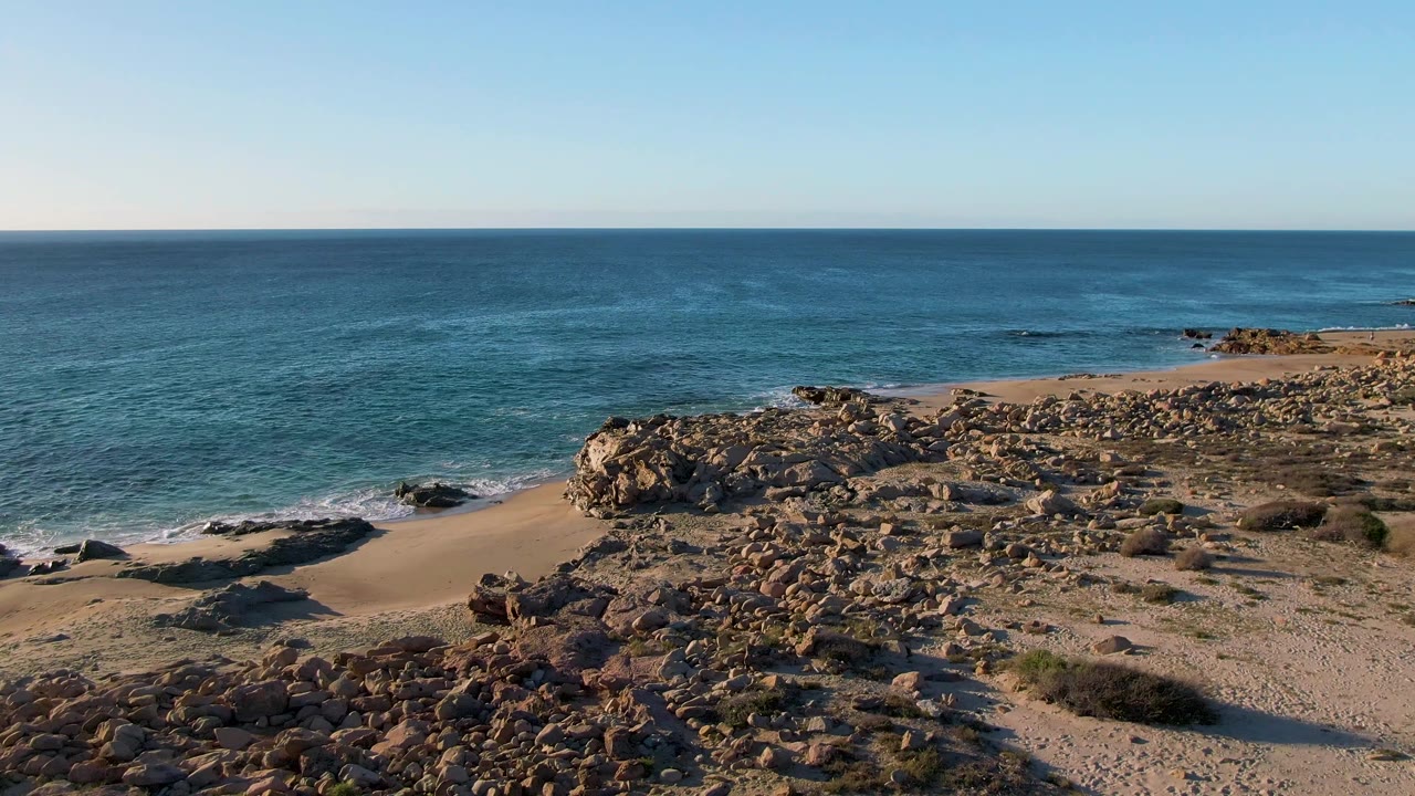 Aerial view of rocky beach, sand, blue ocean, and clear sky.