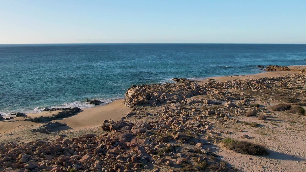 Aerial view one second in at Buzzards Beach East Cape BCS Mexico, showing a small sandy cove between rock outcrops with gentle white surf and no visible people.