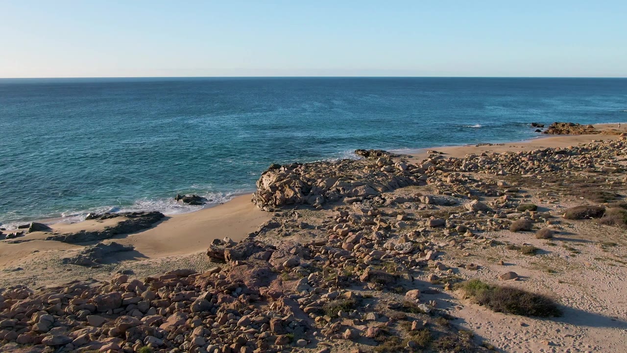 Mid-clip aerial frame over Buzzards Beach East Cape BCS Mexico with the ocean horizon high in frame, rocks across the foreground, sand along the left shoreline, and no visible people.