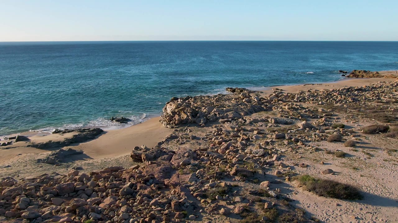 Aerial frame at 0:03 over Buzzards Beach East Cape BCS Mexico, with darker rock shadows in the foreground, a sandy beach strip at left, blue sea behind, and no visible people.