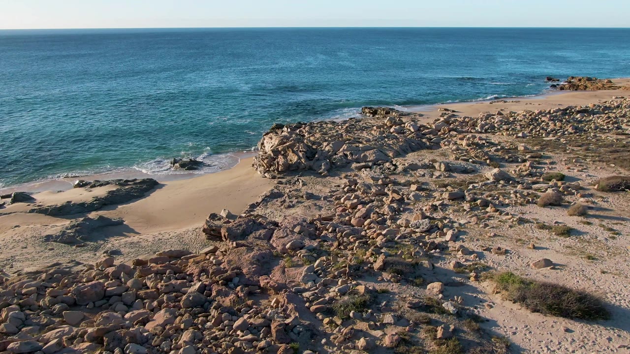 Opening frame of Buzzards Beach in East Cape, BCS Mexico, with turquoise water, pale sand, boulder-strewn shore, sparse scrub, and no visible people.