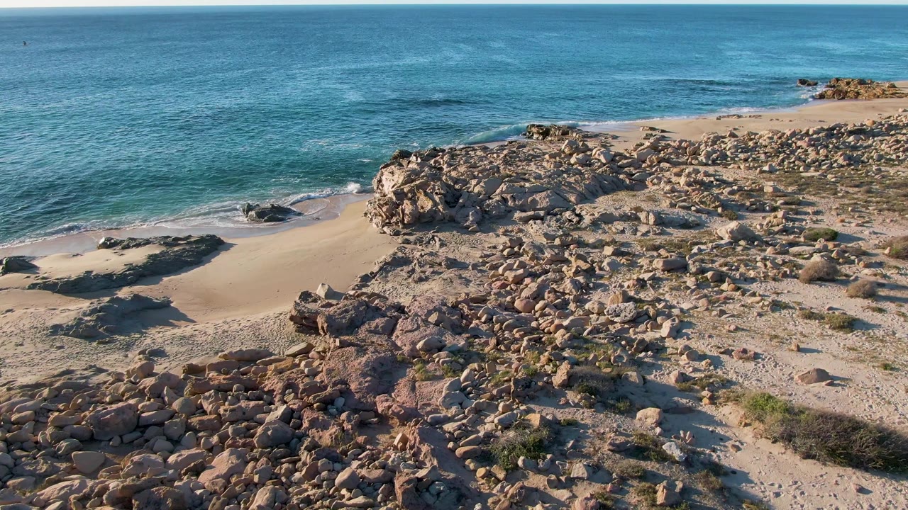 Elevated frame over Buzzards Beach in East Cape, BCS Mexico, with the boulder field filling the lower frame, calm blue ocean beyond, thin surf at the sand, and no visible people.